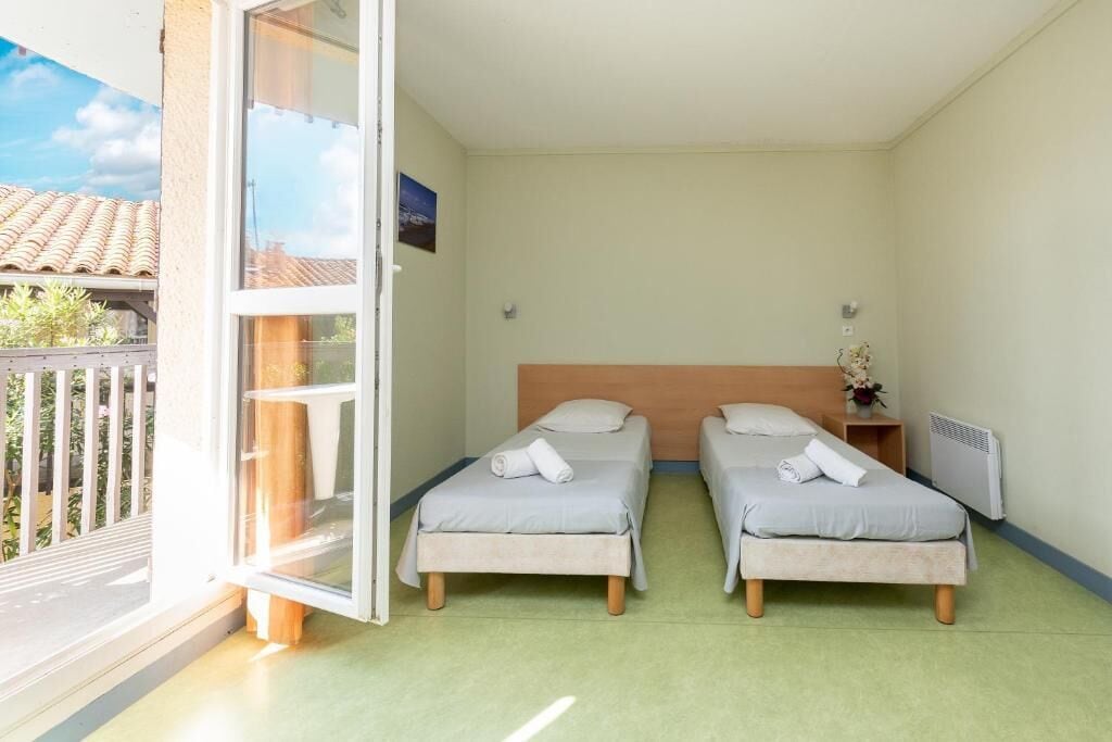 A room with two single beds, a wooden headboard, a small bedside table with a vase, and a window with a view of the outside.