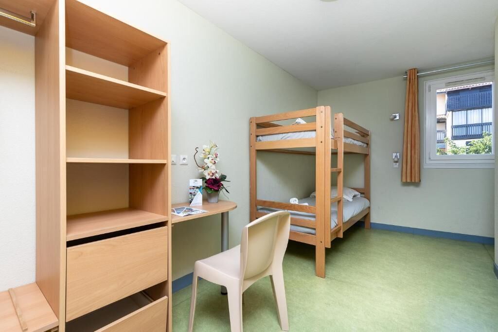 A bunk bed room with a desk and chair, a wooden wardrobe, and a window with a view of buildings.
