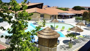 A pool area with lounge chairs, a thatched-roof structure, and a fenced pool. - Village Vacances Le Junka (Vieux-Boucau-les-Bains)
