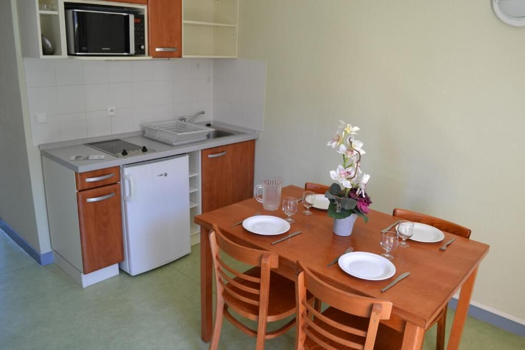 A compact kitchen with a microwave, sink, and a small refrigerator. There is a dining table set with plates, cutlery, and a vase with flowers.