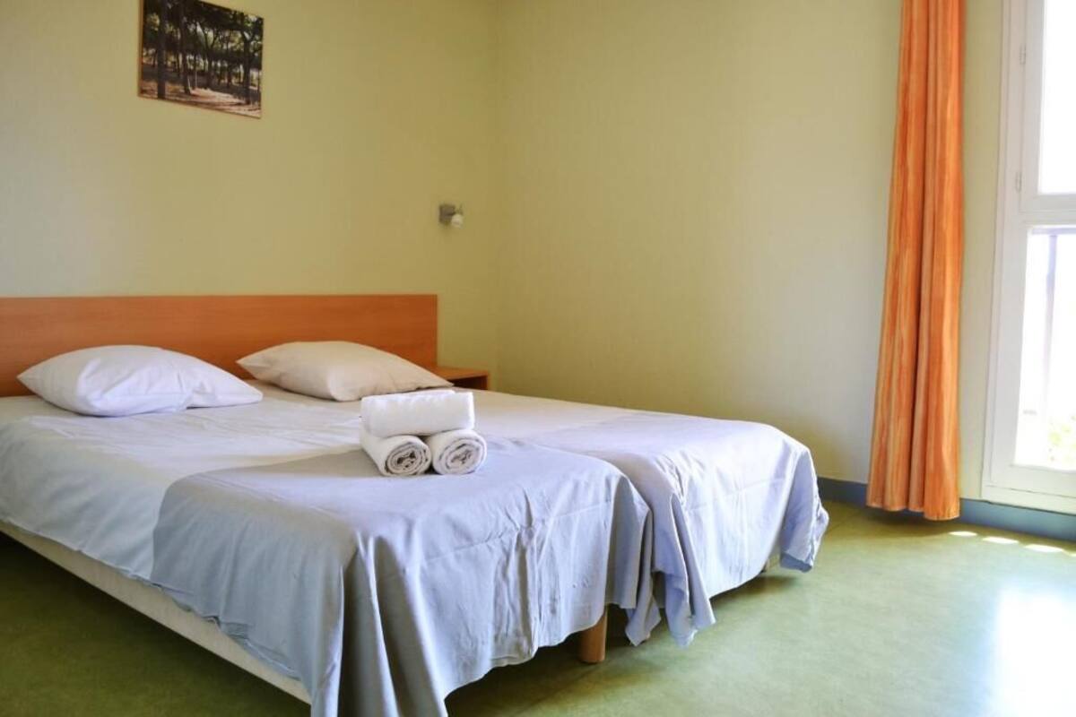 A room with two single beds, a wooden headboard, a small table, and a window with orange curtains.
