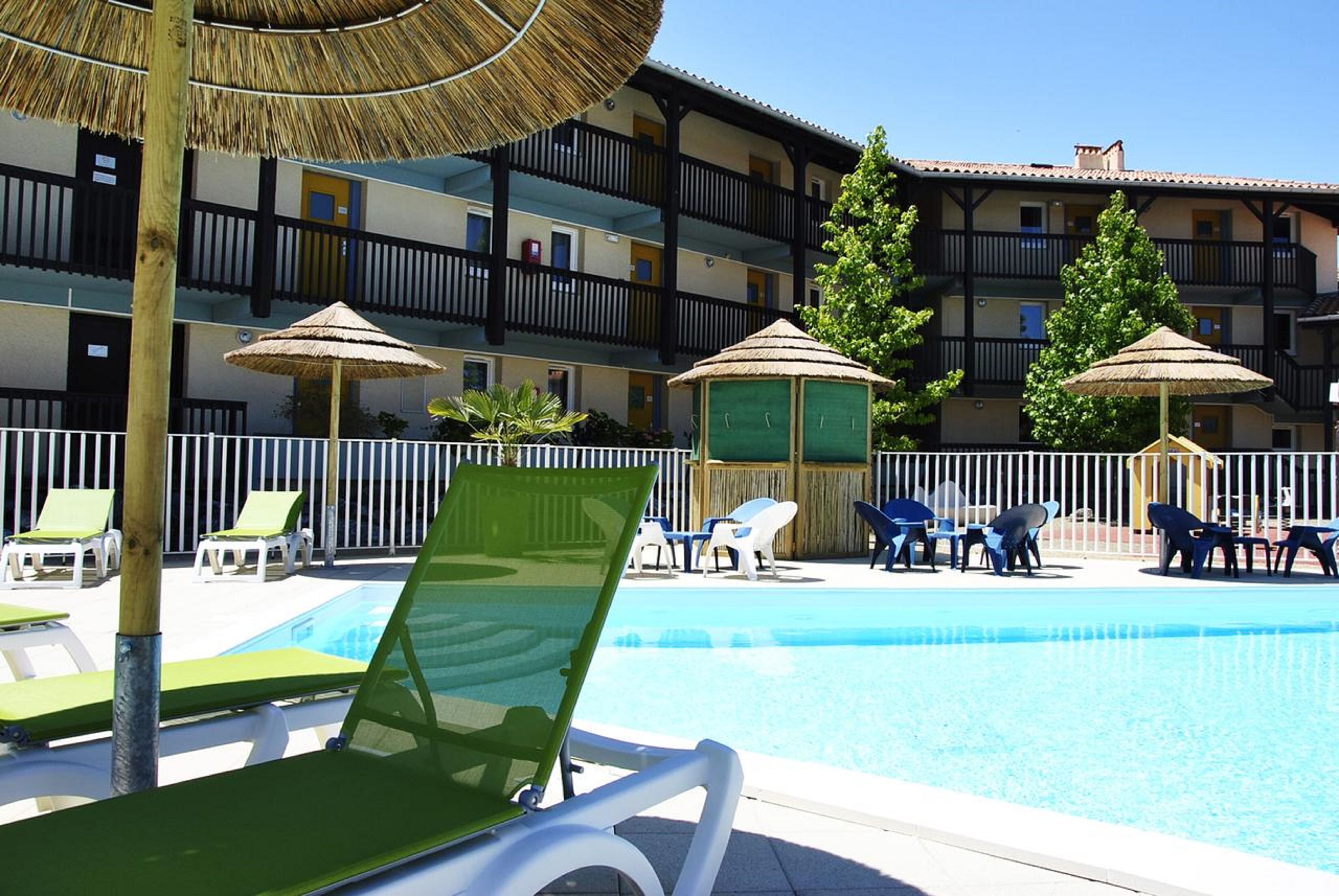 A pool area with lounge chairs and umbrellas, a building with balconies, and a clear blue sky.