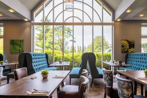 Een restaurant met hoge plafonds, grote ramen en uitzicht op groen.