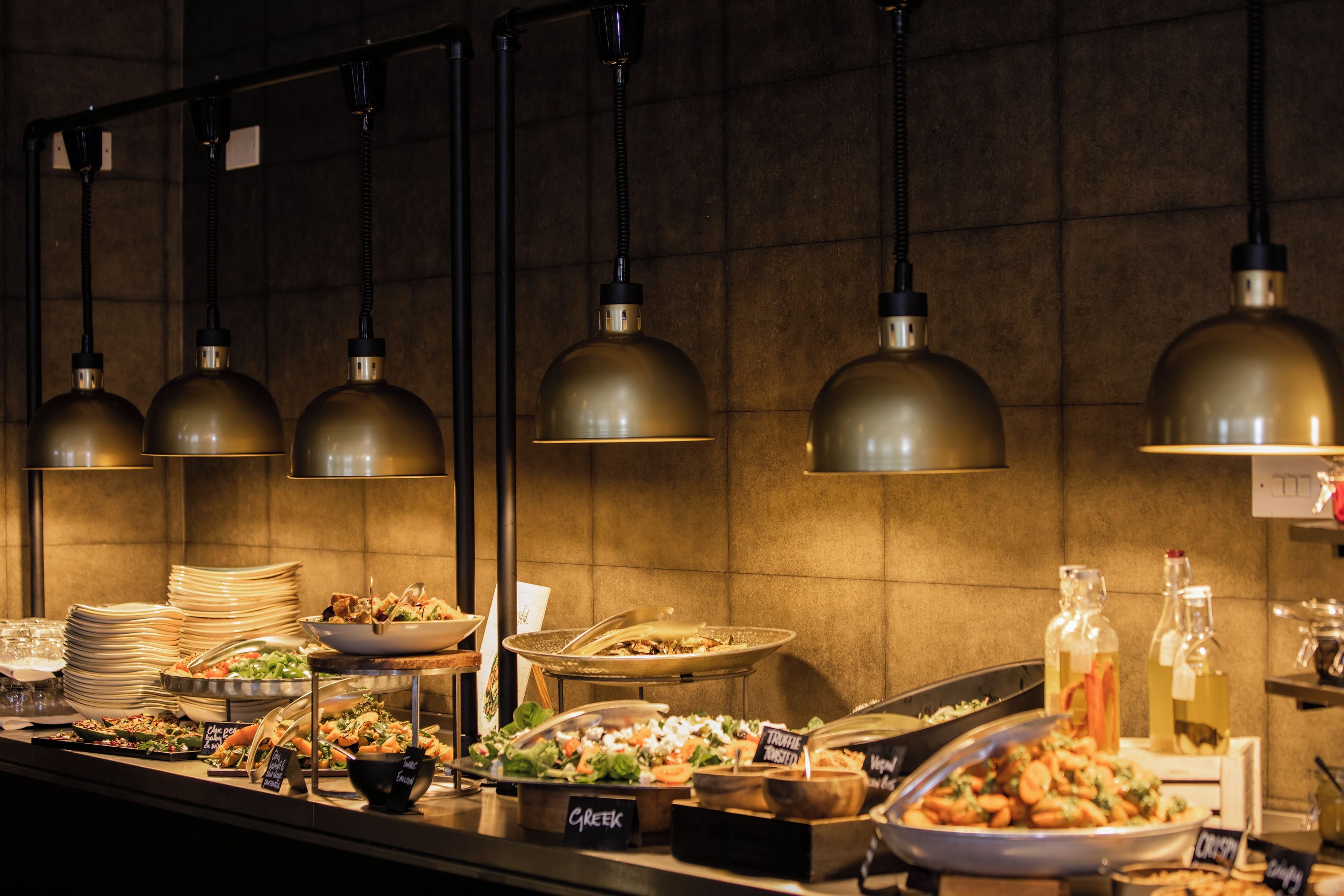 Montaje de bufé con varias estaciones de comida, que incluyen opciones griegas y veganas, bajo iluminación colgante de estilo industrial.
