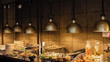 A buffet setup with various food stations, including Greek and vegan options, under industrial-style pendant lighting.