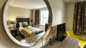 Een hotelkamer met een grote spiegel, een bed met een gestreept dekbed, een nachtkastje met een lamp, een stoel en een bureau met een laptop.
