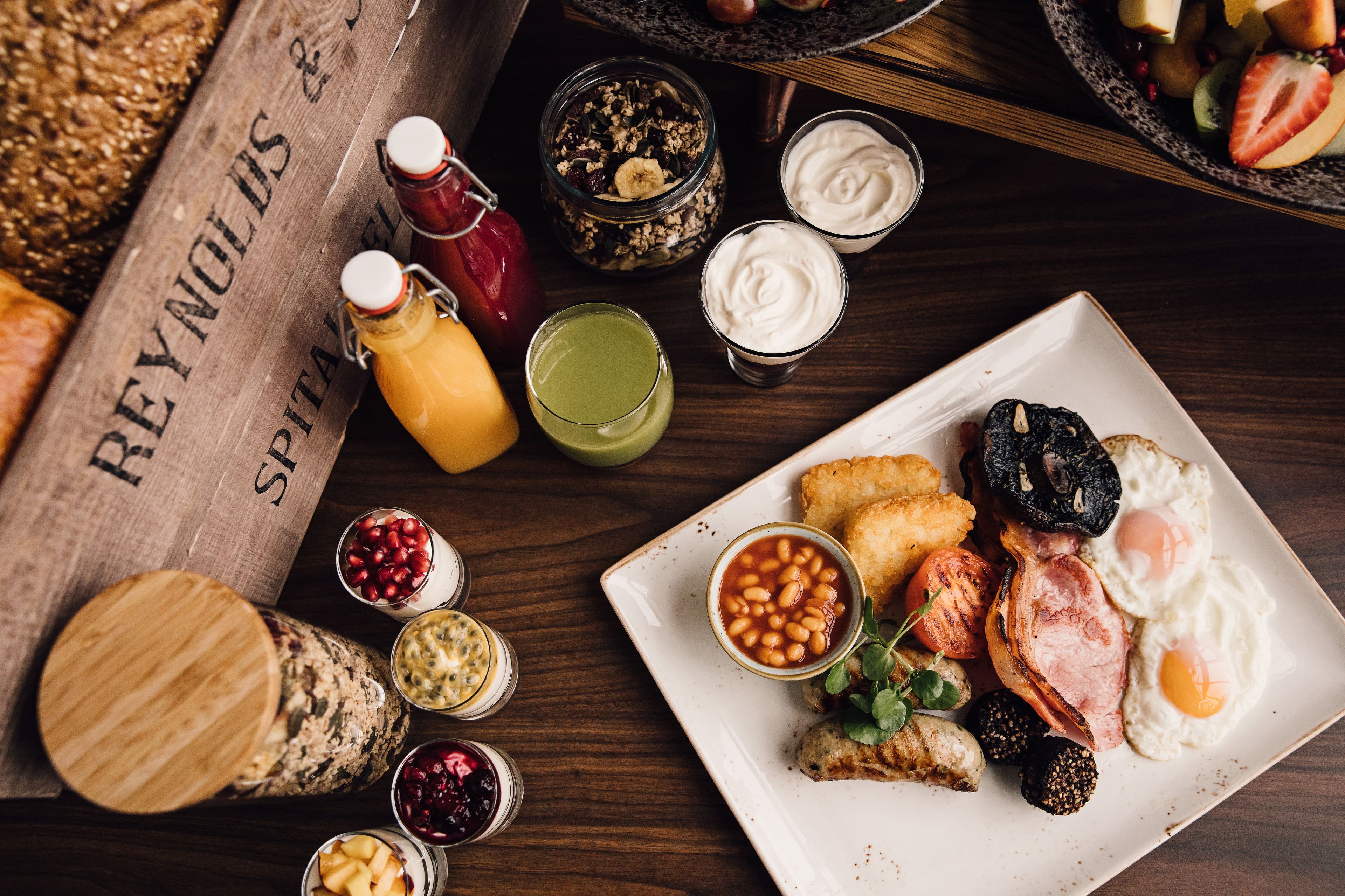 A breakfast plate with eggs, bacon, sausages, and beans, surrounded by various condiments and drinks.