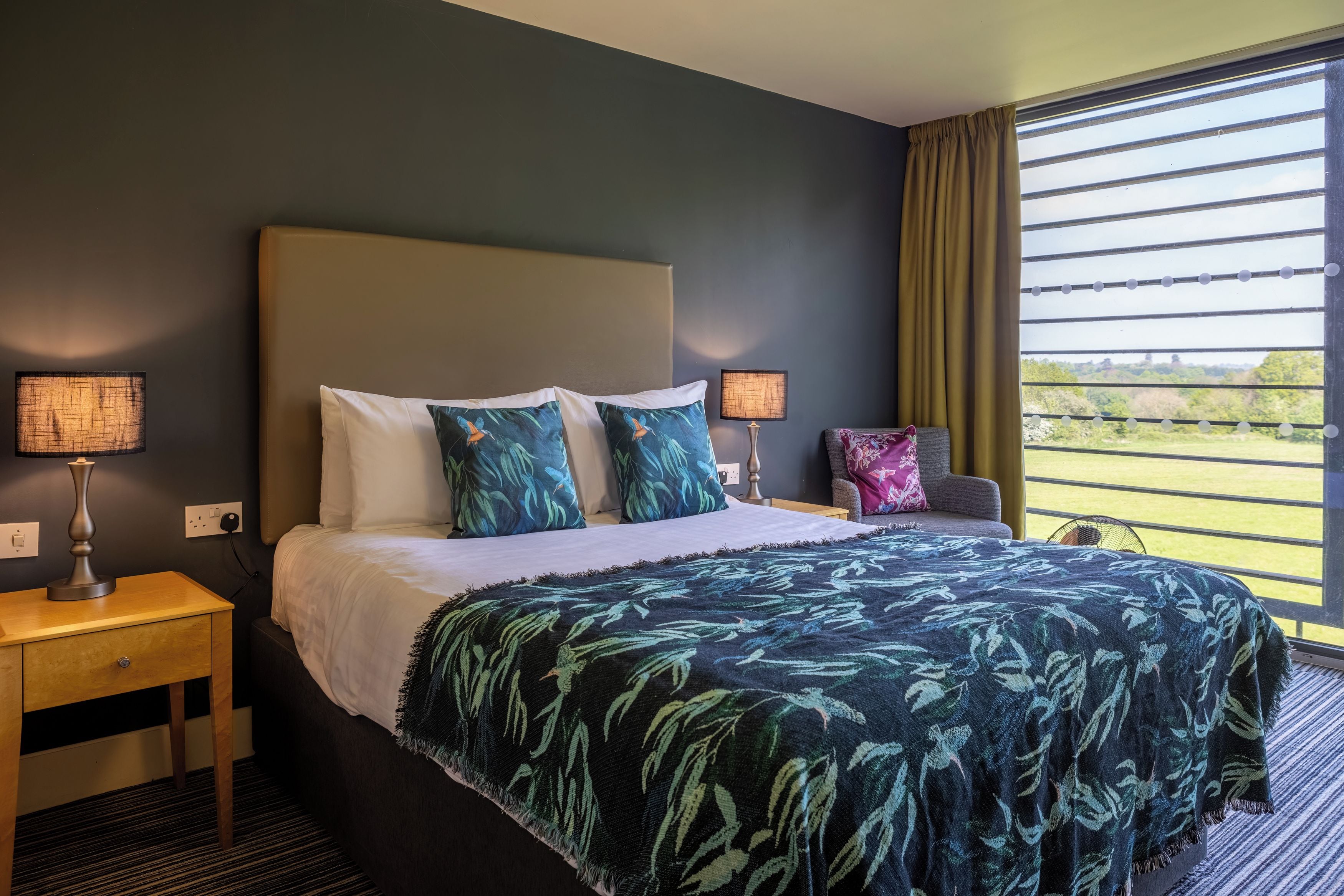 Een hotelkamer met een bed, twee nachtkastjes, een lamp, een stoel en een raam met jaloezieën.