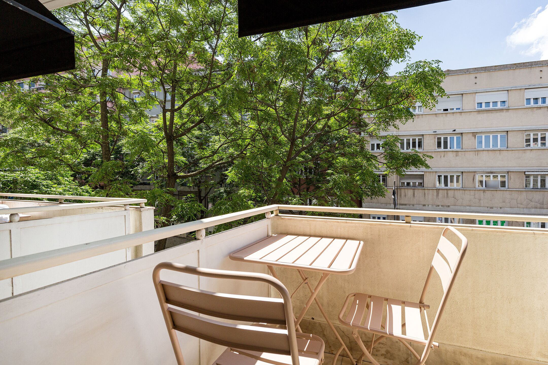 A balcony with two metal chairs and a small table, surrounded by trees and a building in the background.