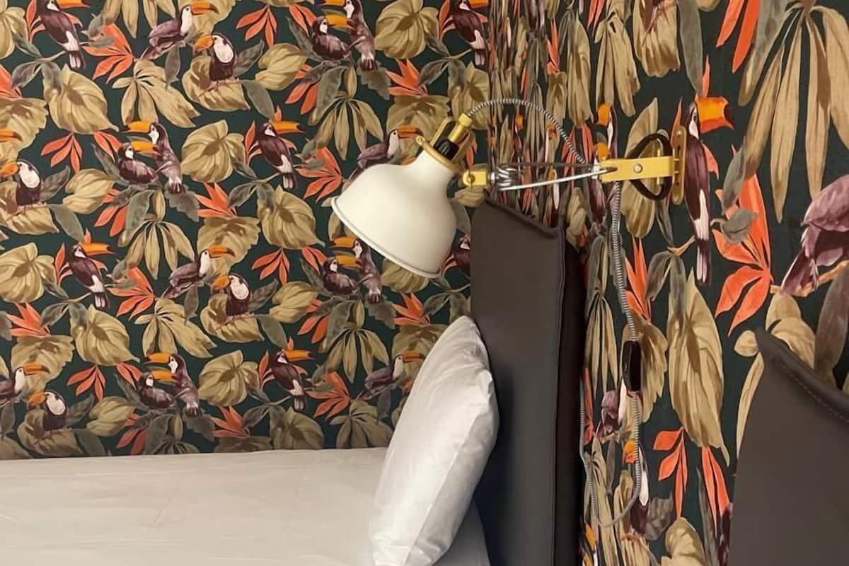 Una habitación con una cama, una mesita de noche y una lámpara. Las paredes presentan un estampado floral.