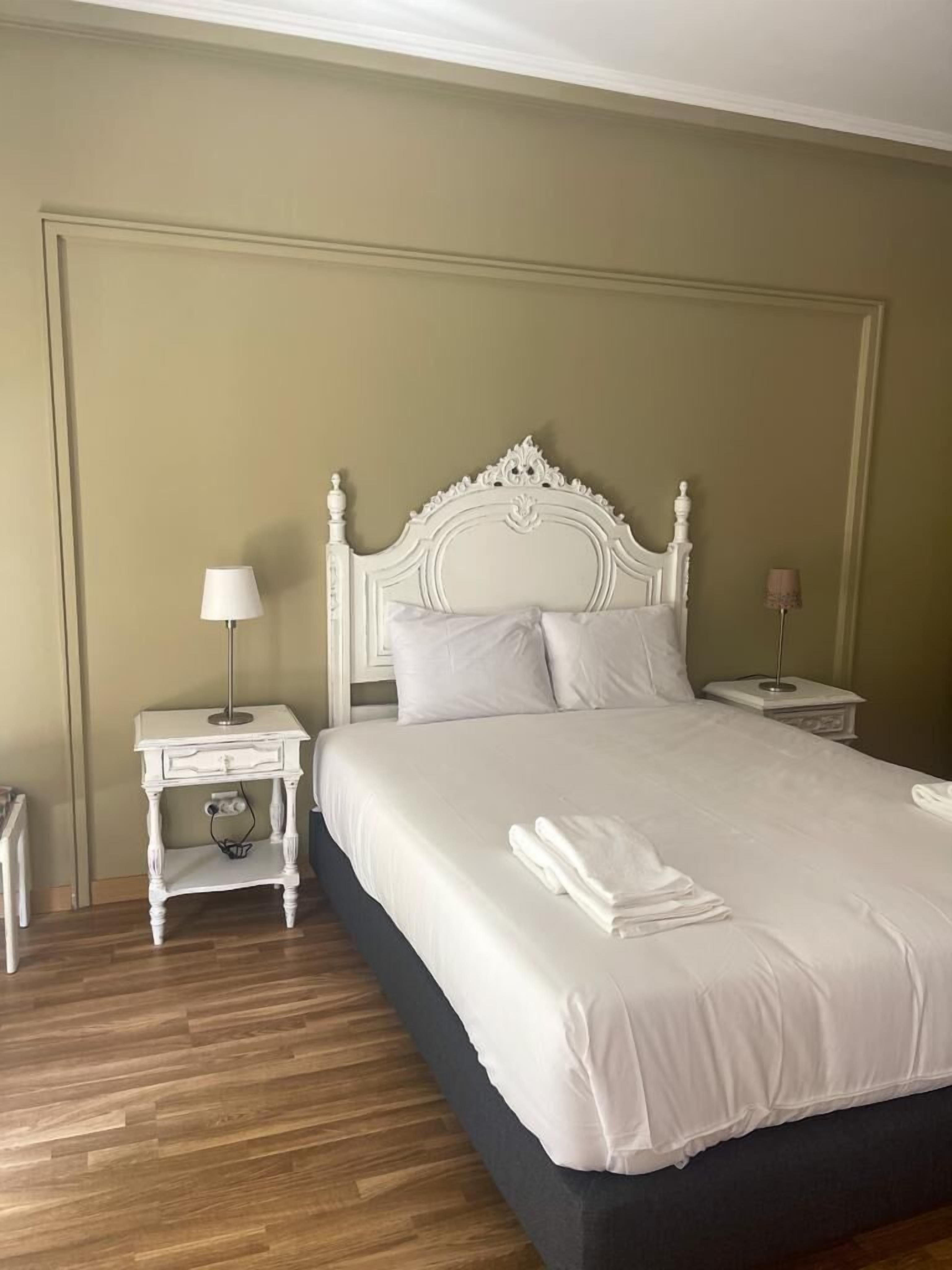 Cama bem arrumada com roupa de cama branca, cabeceira branca e duas mesas de cabeceira com abajures.