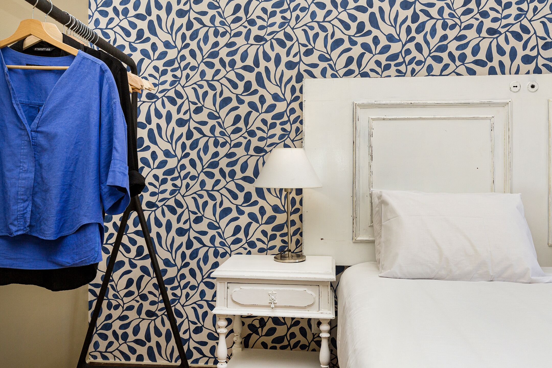 Een slaapkamer met behang met een patroon, een witte nachtkastje met een lamp, een wit bed, en een blauw shirt hangend aan een houten kapstok.