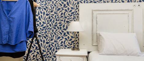 Een slaapkamer met behang met een patroon, een witte nachtkastje met een lamp, een wit bed, en een blauw shirt hangend aan een houten kapstok.