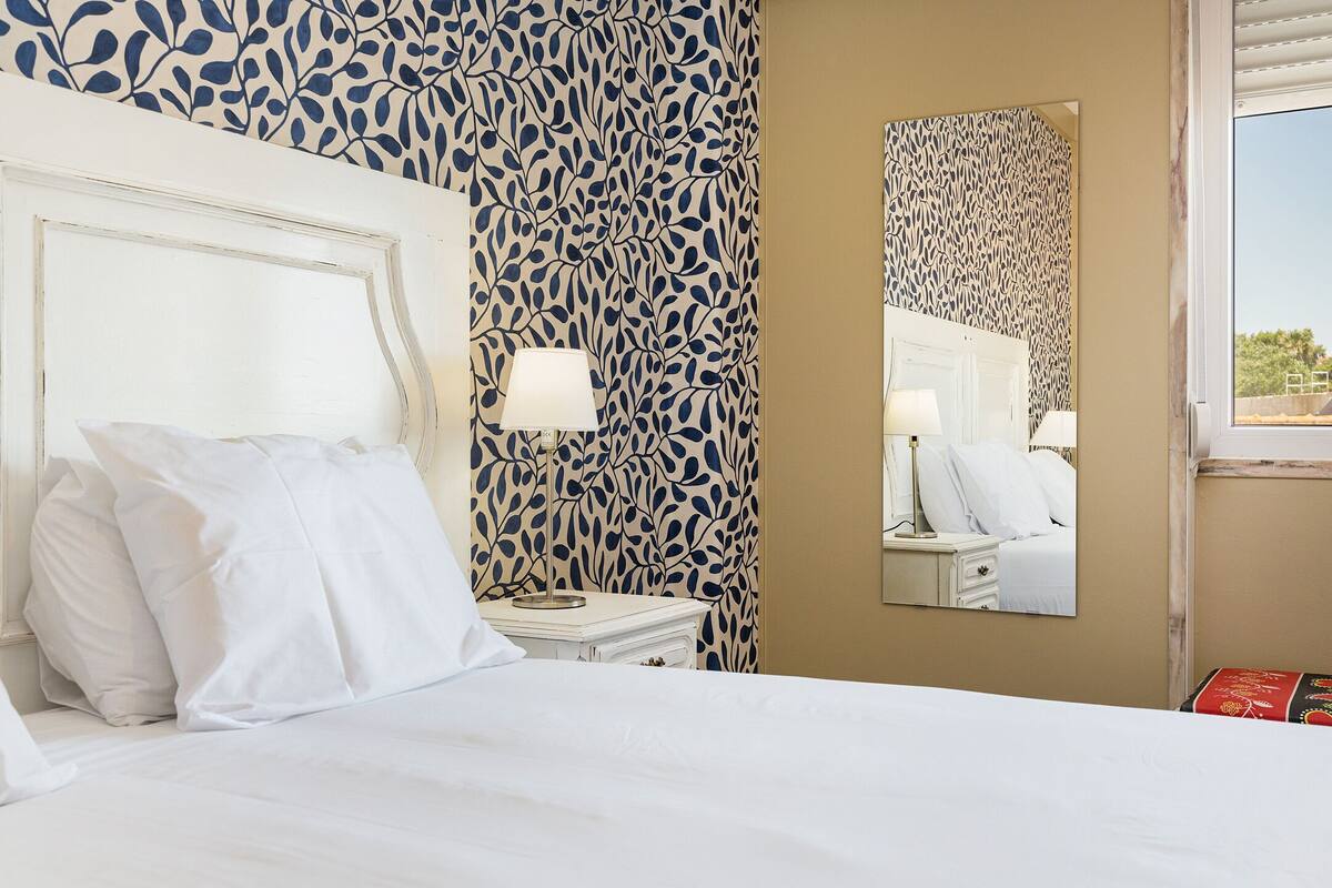 Habitación de hotel con cama, ropa de cama blanca, mesita de noche con lámpara, espejo y un papel tapiz estampado.