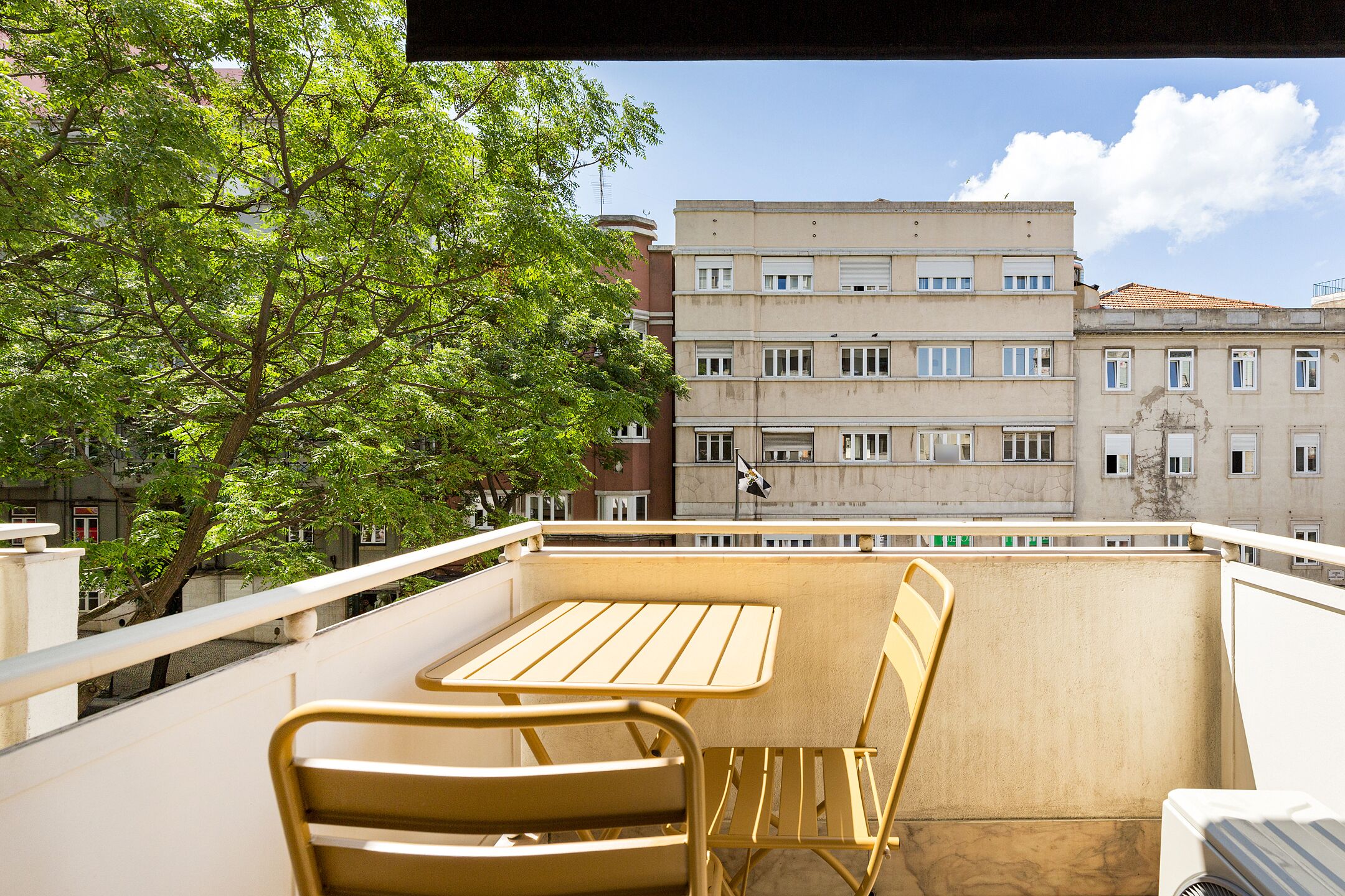 A balcony with a table and chairs, overlooking a tree and buildings.