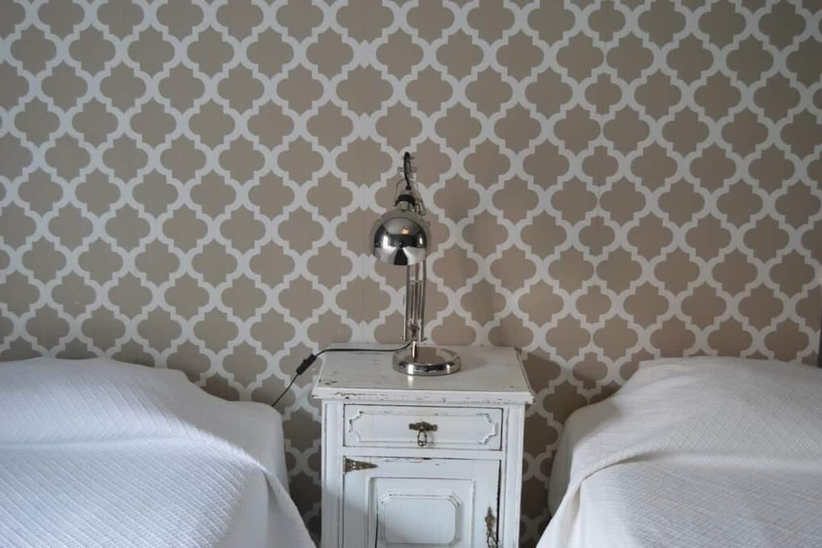 Una habitación con dos camas, una mesita de noche blanca con lámpara y papel tapiz estampado.