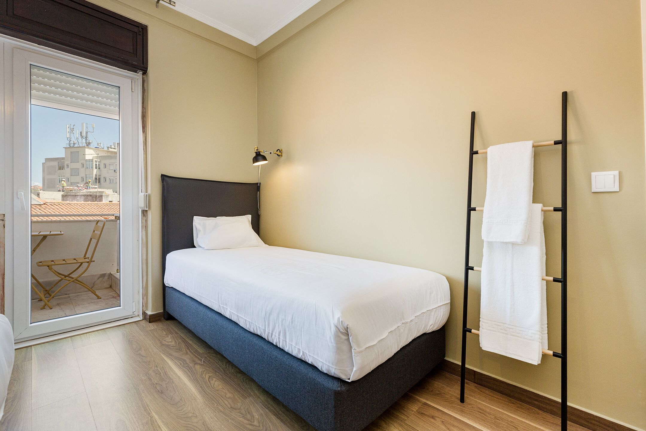 A bedroom with a bed, a nightstand, a lamp, a towel rack, and a view of the outside through a sliding door.