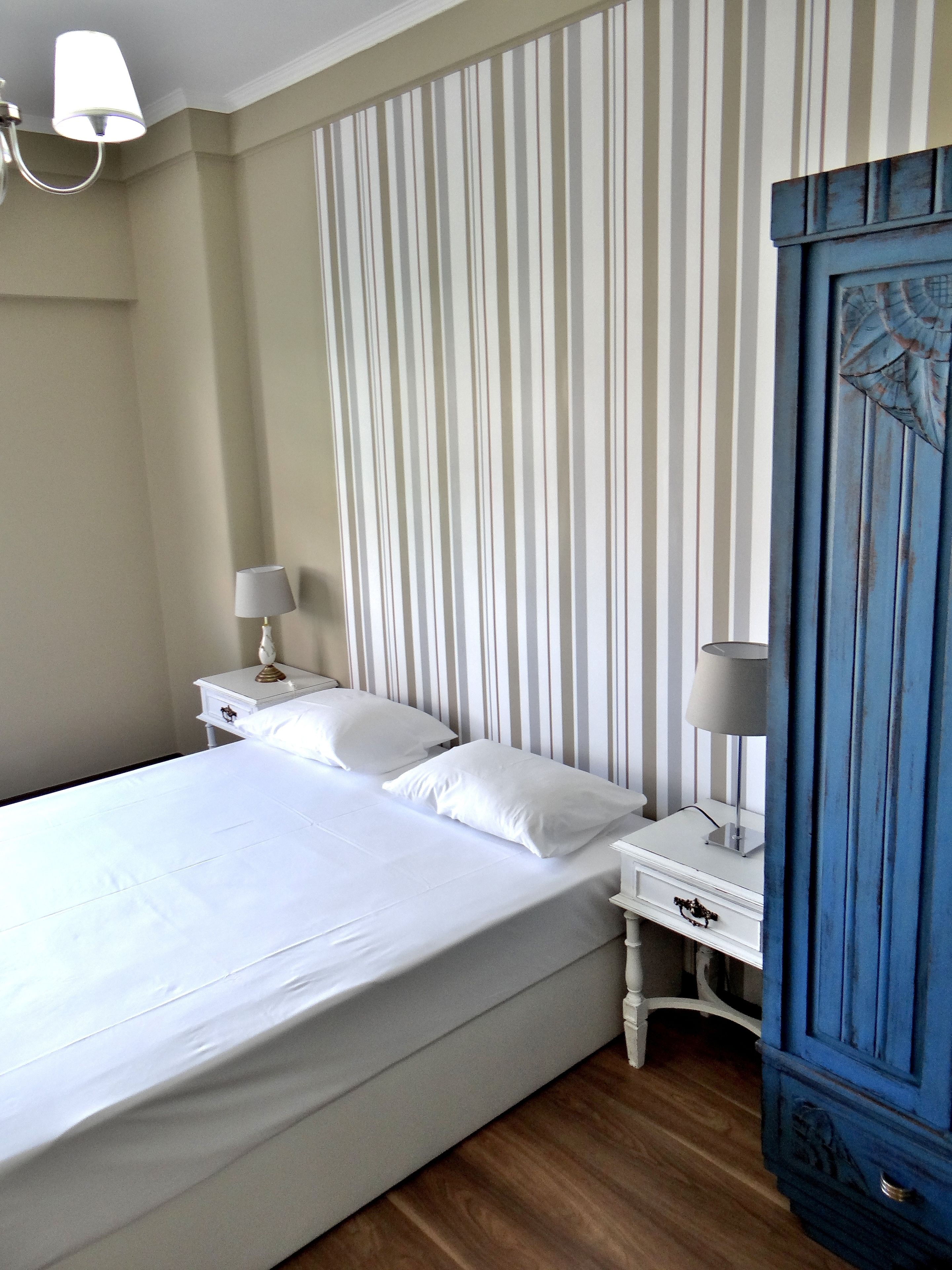 A neatly made bed with white linens in a room with striped wallpaper, a wooden floor, and a blue cabinet.