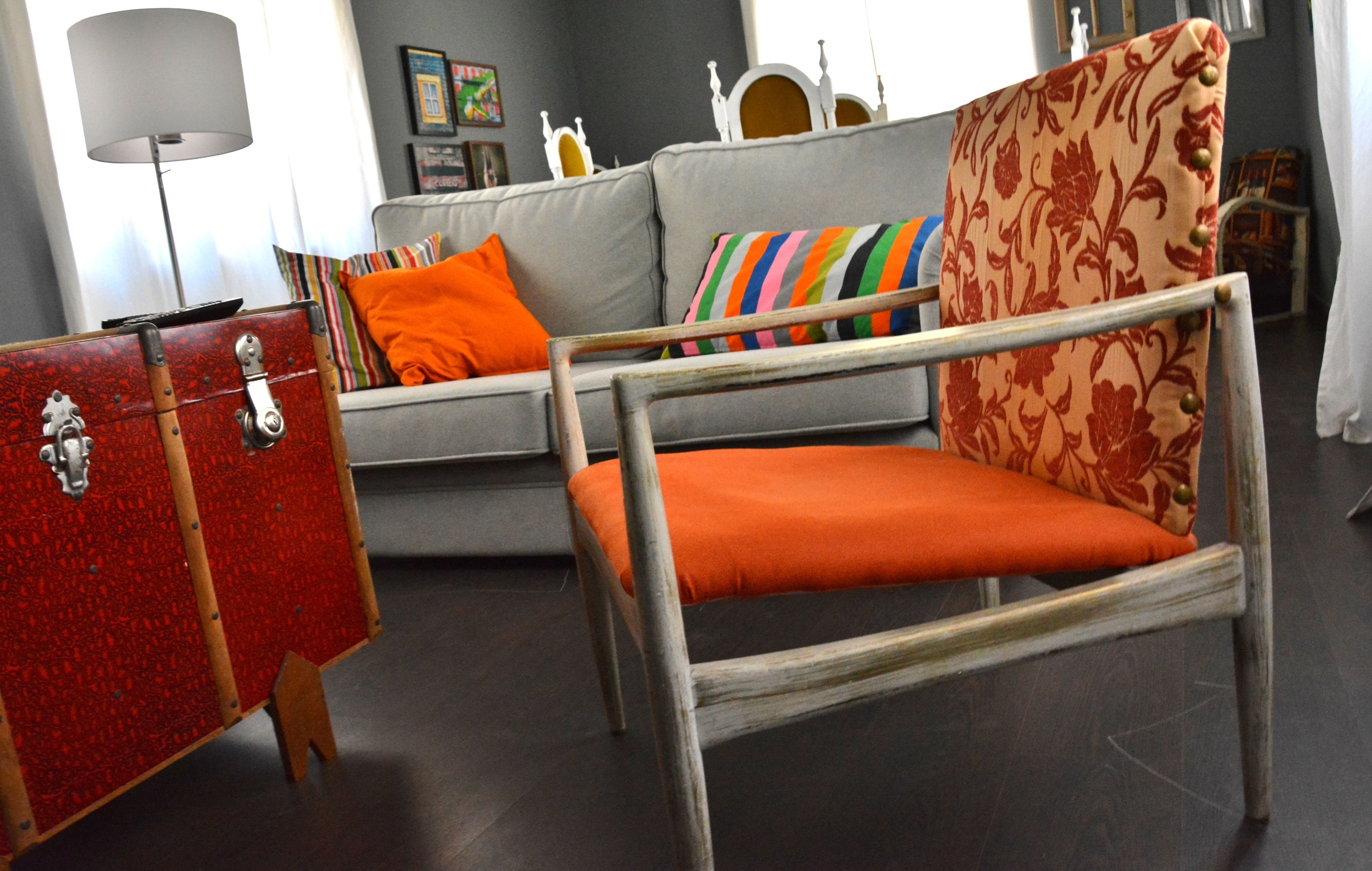 Una habitación con un sofá gris, almohadones multicolores a rayas, una maleta roja y una silla de madera con cojines naranjas.