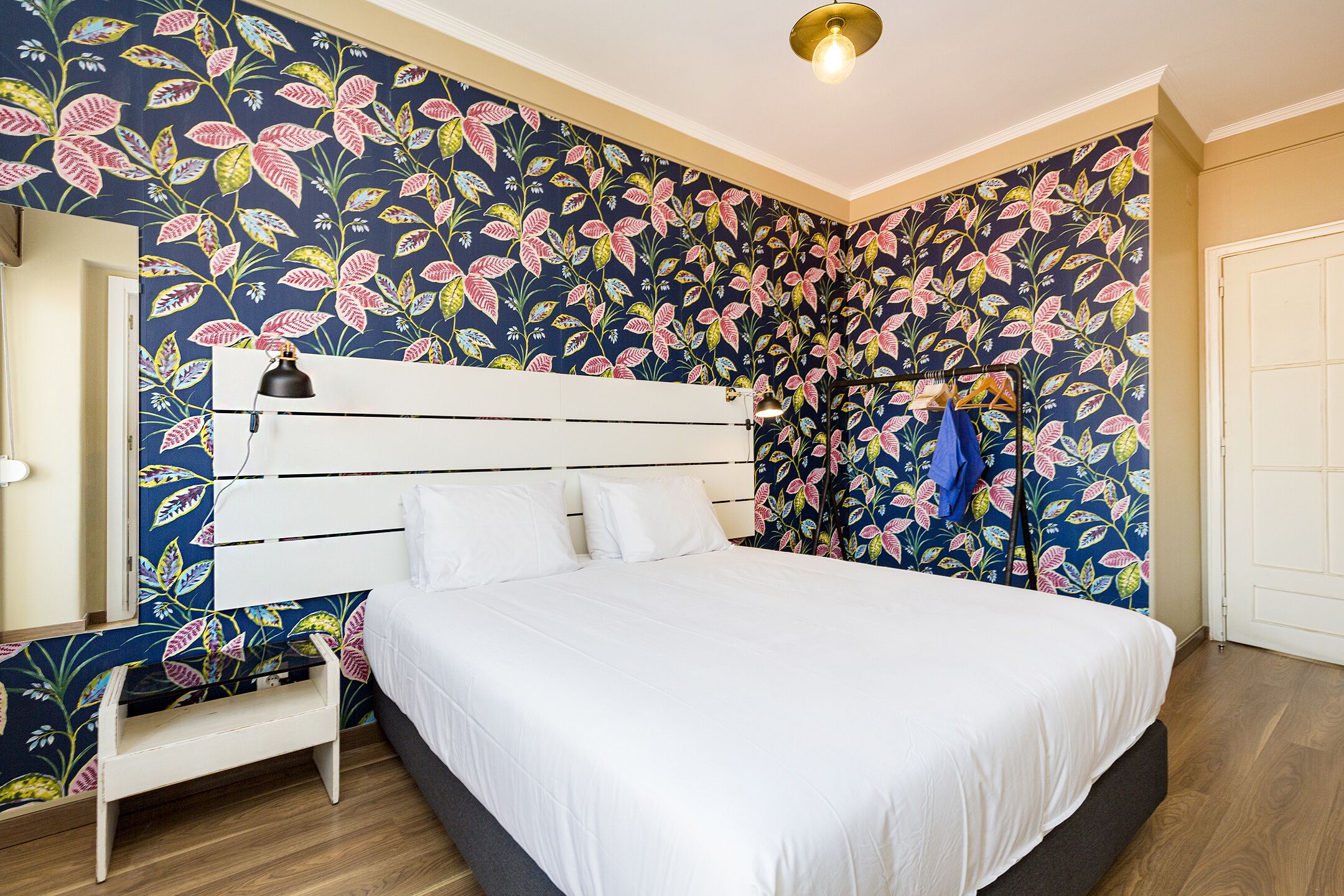 A bedroom with a bed, a white nightstand, a wall-mounted lamp, and a door.