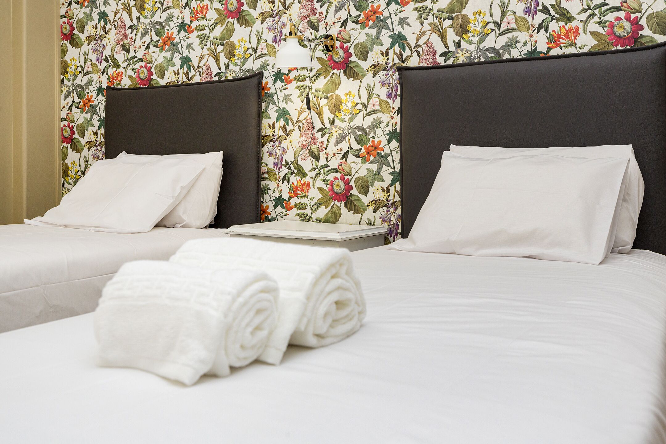 Two twin beds with white linens and pillows, a floral wallpaper, and a small bedside table with a lamp.