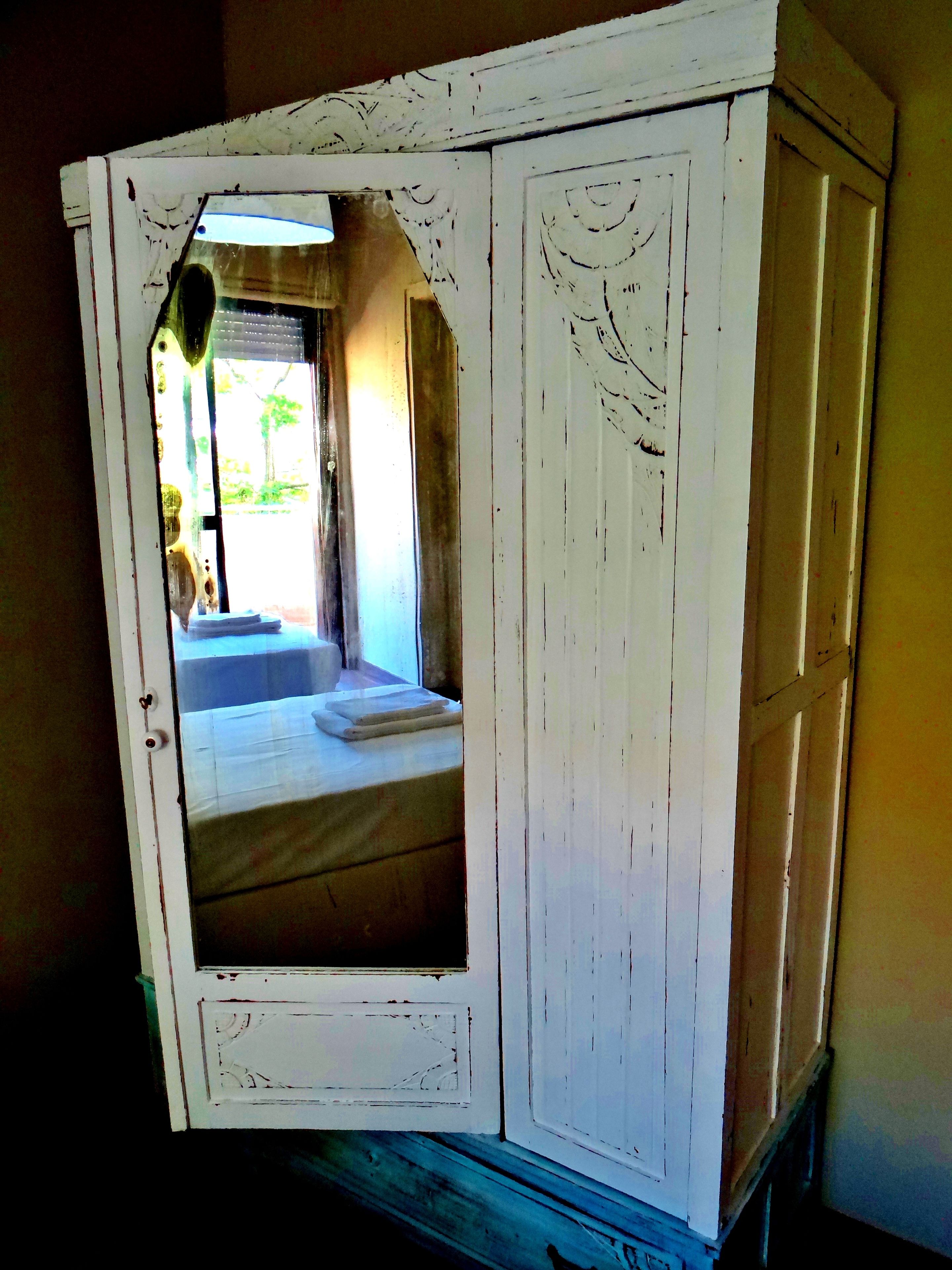 A white, rustic cabinet with a small window, revealing a glimpse of a room with a bed and a window.