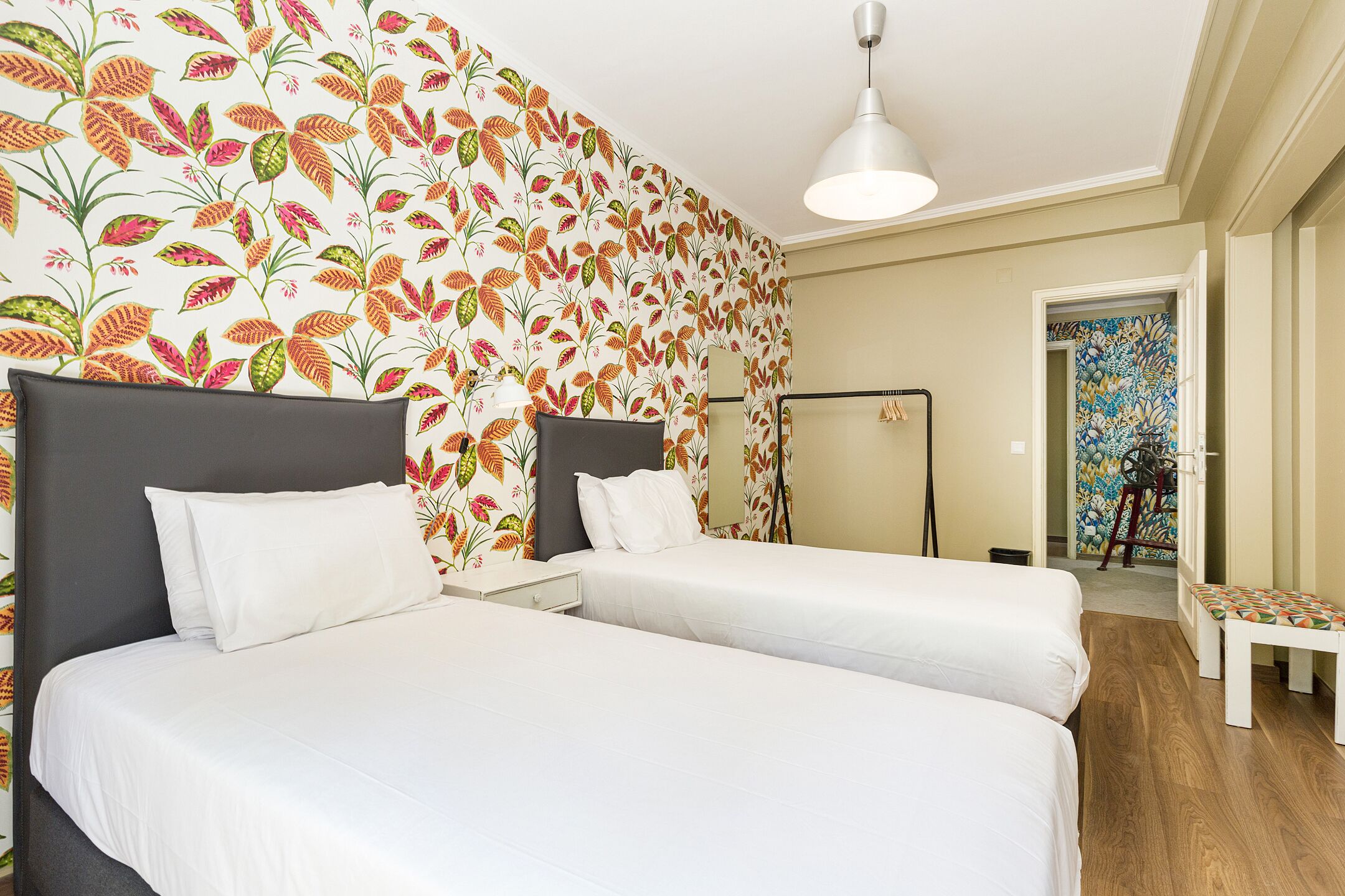 Habitación de hotel con dos camas, una mesita de noche, una lámpara y un papel tapiz floral de colores.