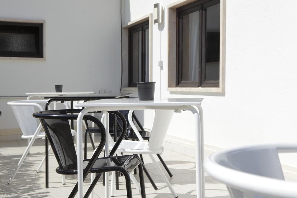 A patio area with white and black chairs and tables, a small trash bin, and a window with blinds.