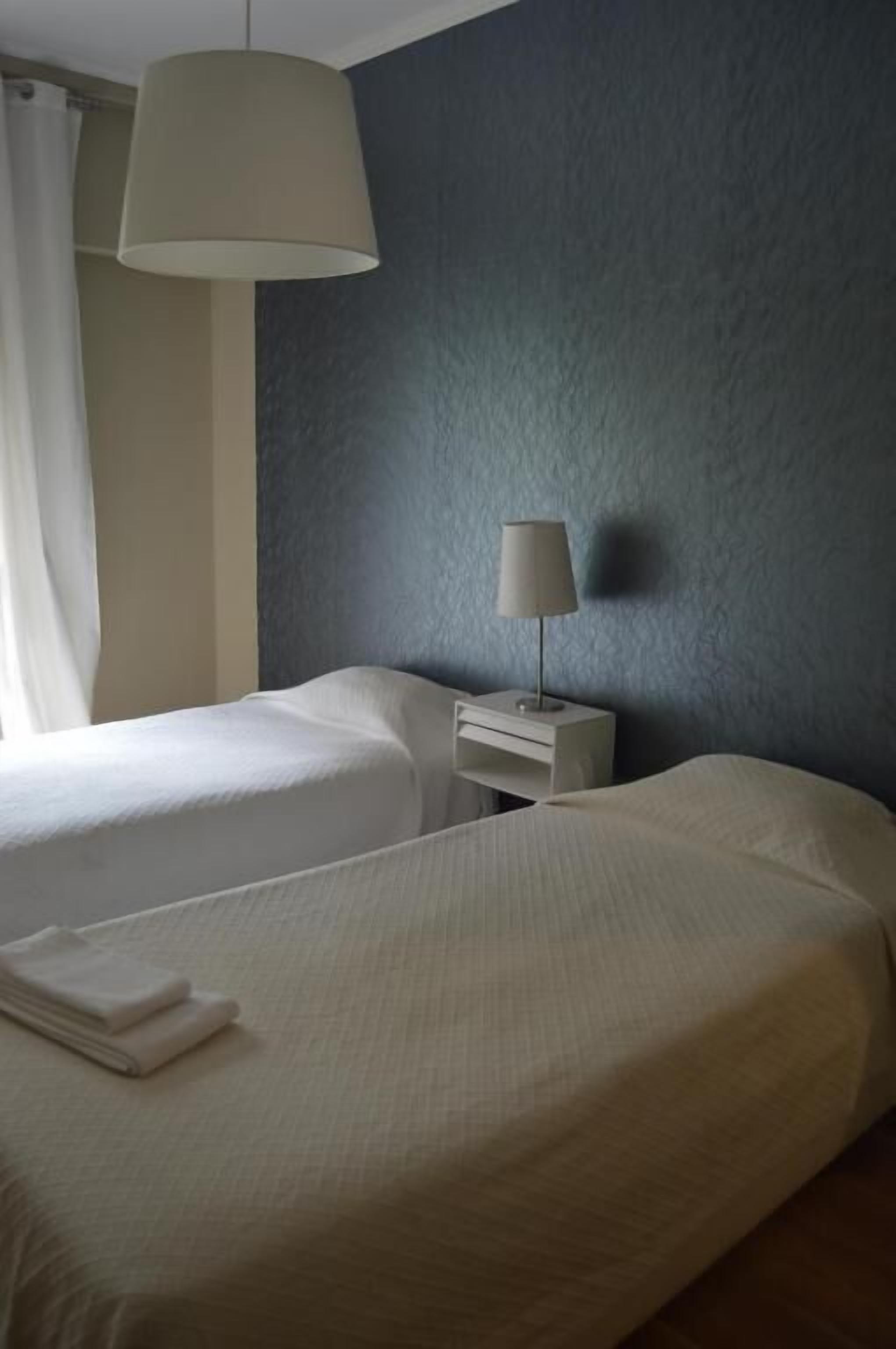 A hotel room with two beds, a nightstand with a lamp, and a large window with curtains.