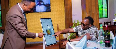 A man in a suit holding a tablet displaying a menu at a restaurant table with a wine bottle and glasses.