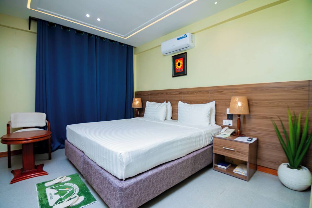 A hotel room with a bed, bedside tables, a chair, a desk, a lamp, a plant, and an air conditioning unit.