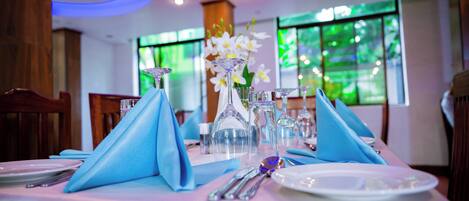 A dining table set with blue napkins, clear glassware, and white plates, with a vase of white flowers in the center.