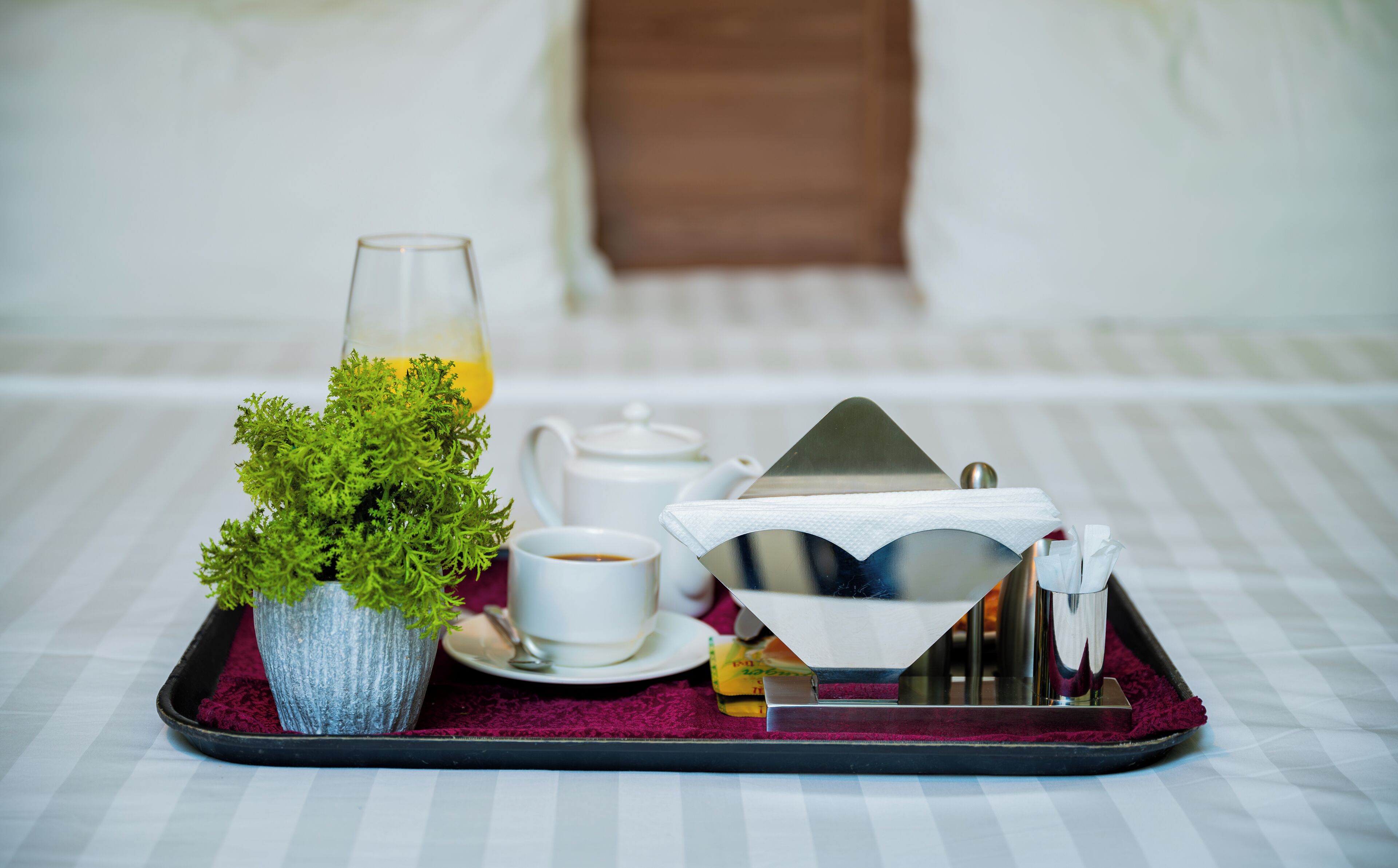 Ein Tablett mit einem Blumentopf, einem Glas gelber Flüssigkeit, einer weißen Teekanne, einer weißen Tasse und einem silbernen Tablettständer steht auf einem Bettlein mit Streifenmuster.