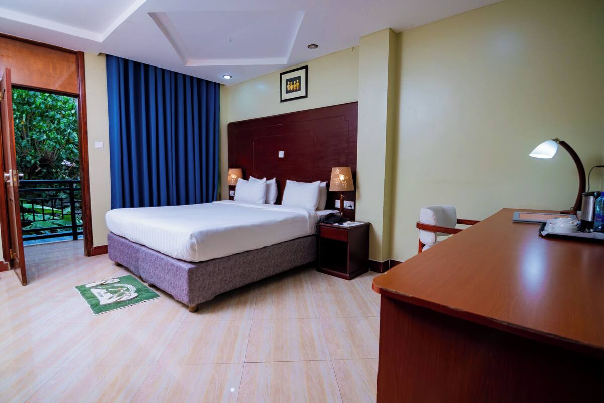 A hotel room with a bed, bedside tables, a desk, and a view of greenery outside.
