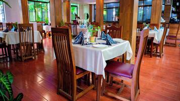A restaurant with wooden chairs and tables, large windows, and a green plant on the table.