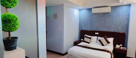 A modern hotel room with a large bed, a nightstand, a potted plant, and an air conditioning unit on the wall.