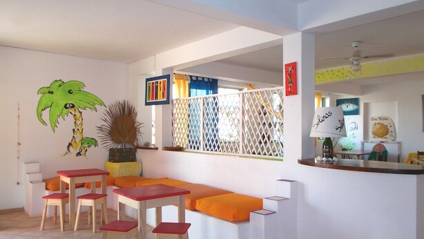 A room with a white wall, a wooden table with red cushions, a bench with orange cushions, and a decorative palm tree mural.
