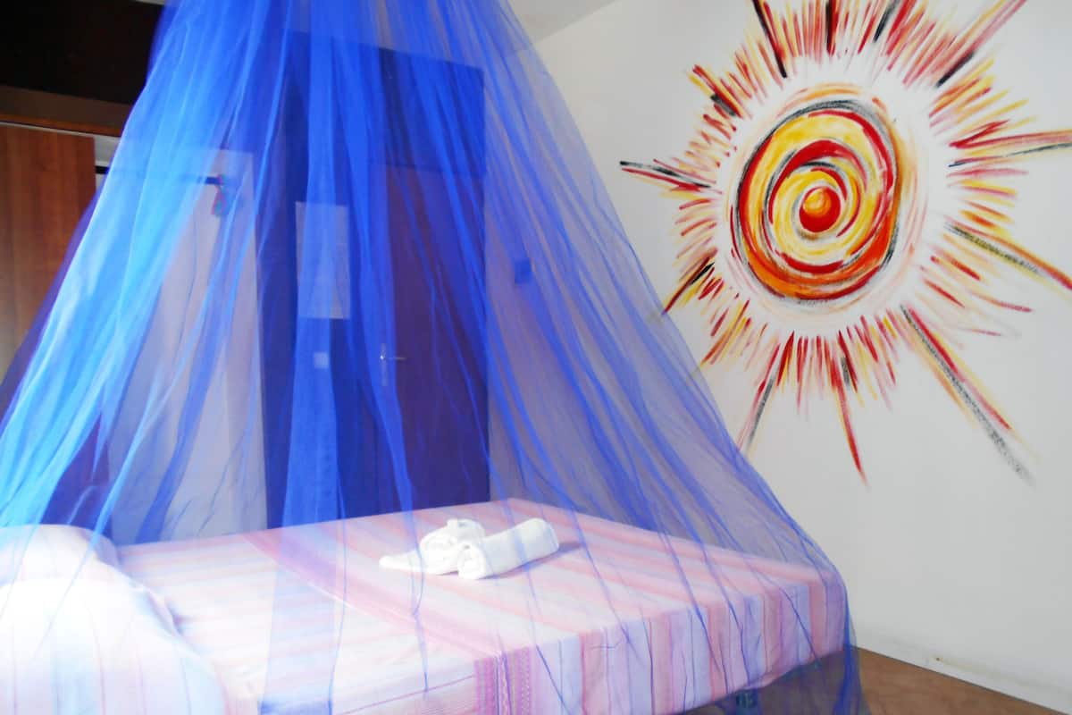 Una cama con dosel azul, un almohadón y una pared con una obra de arte que representa un sol colorido.