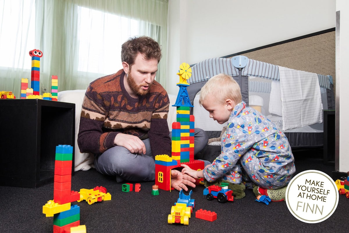A child and an adult playing with building blocks on a carpeted floor in a room with a bed and a window with curtains.