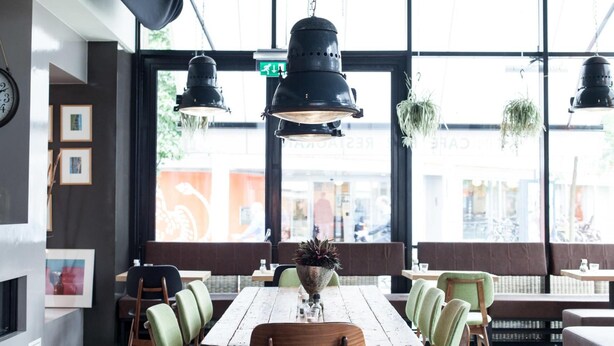 A restaurant interior with a wooden table, green upholstered chairs, hanging pendant lights, and large windows with a view of the street.
