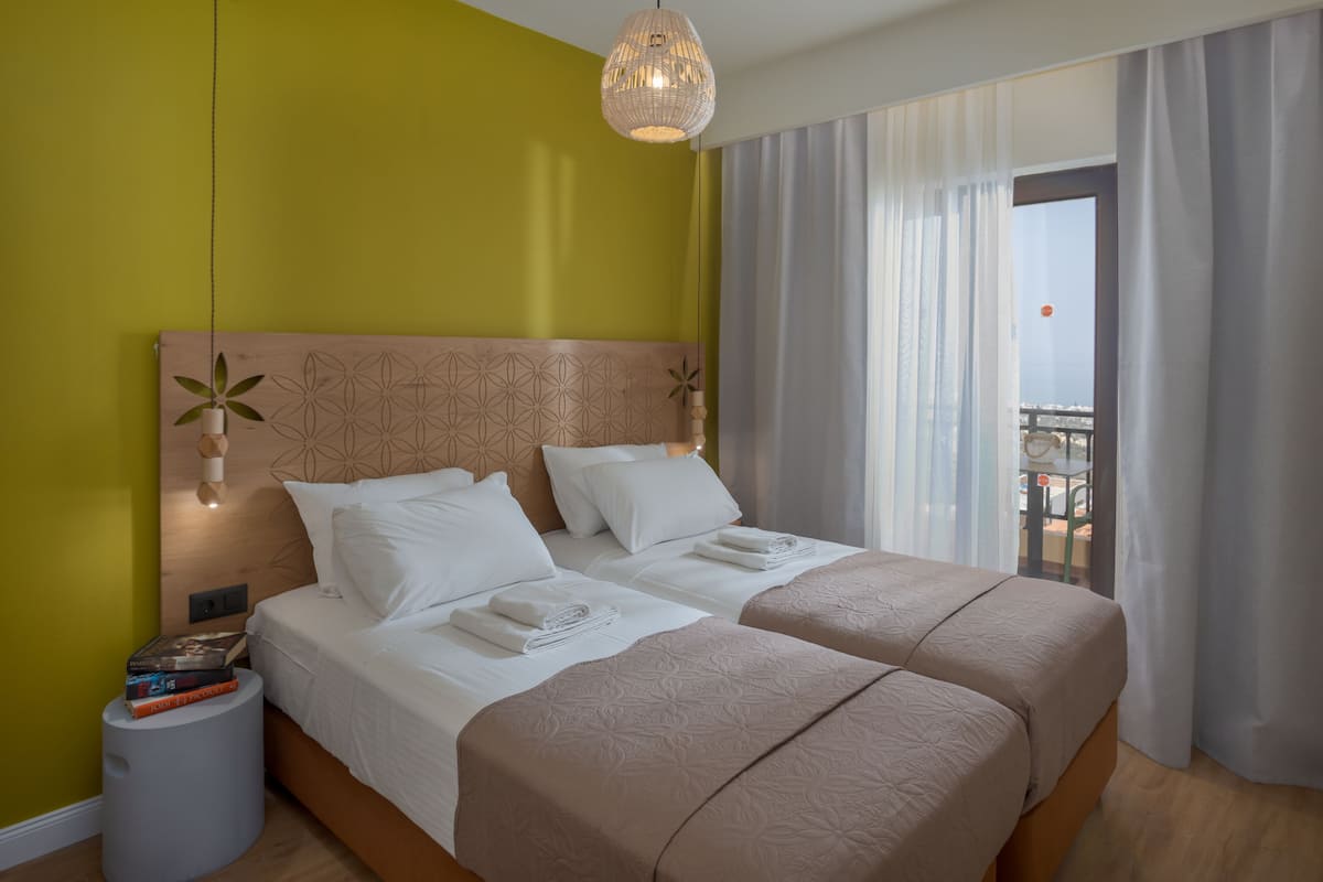 A hotel room with two beds, a large window with curtains, a hanging light fixture, and a small table with books.