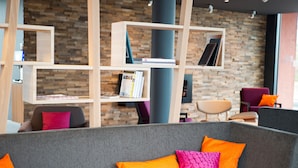 Un área de descanso moderna con un sofá gris, cojines naranja y rosa, y estanterías de madera frente a una pared de ladrillo.