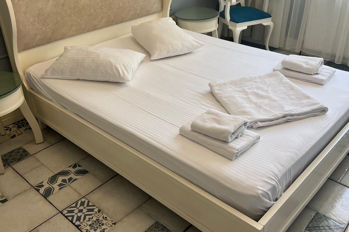 A neatly made bed with pillows and folded towels, a small round table with a chair, and patterned floor tiles.