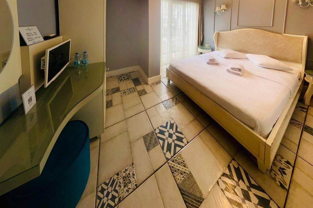 A hotel room with a bed, a desk with a computer, a mirror, and a window with curtains.