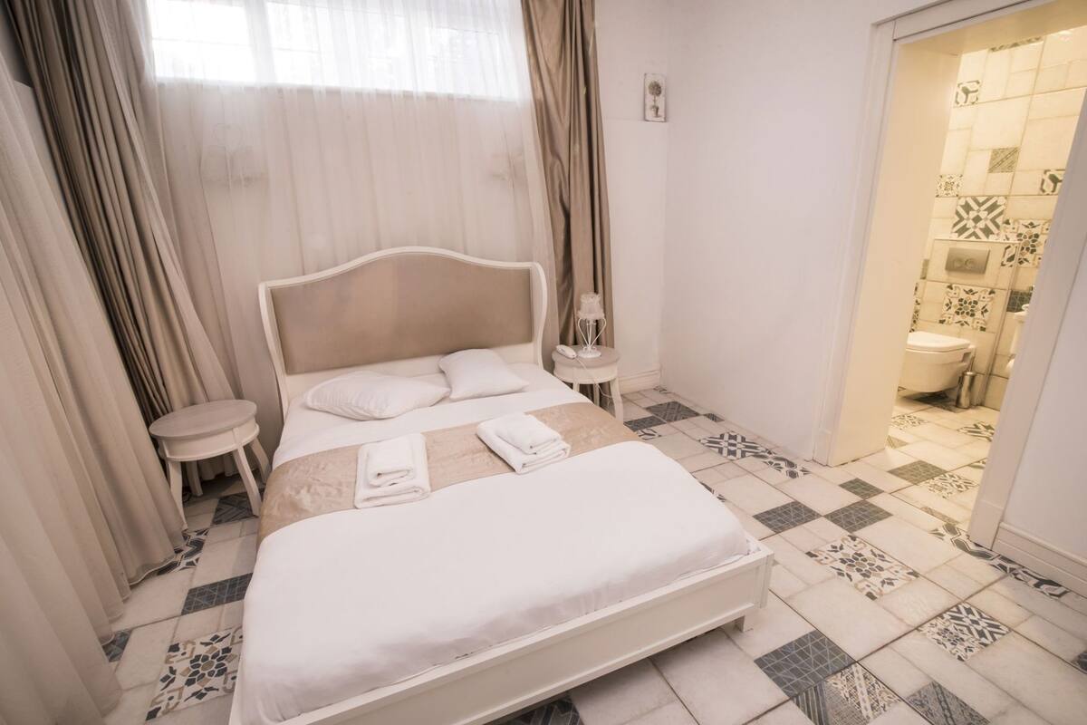 A hotel room with a bed, bedside tables, a nightstand, a window with curtains, and a bathroom visible through an open door.