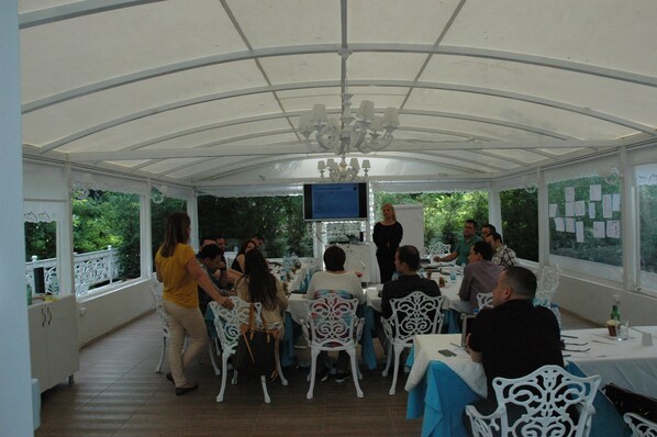 A covered outdoor dining area with white chairs and tables, a chandelier, and a screen displaying information. - Buyukada Cankaya Hotel (Adalar)