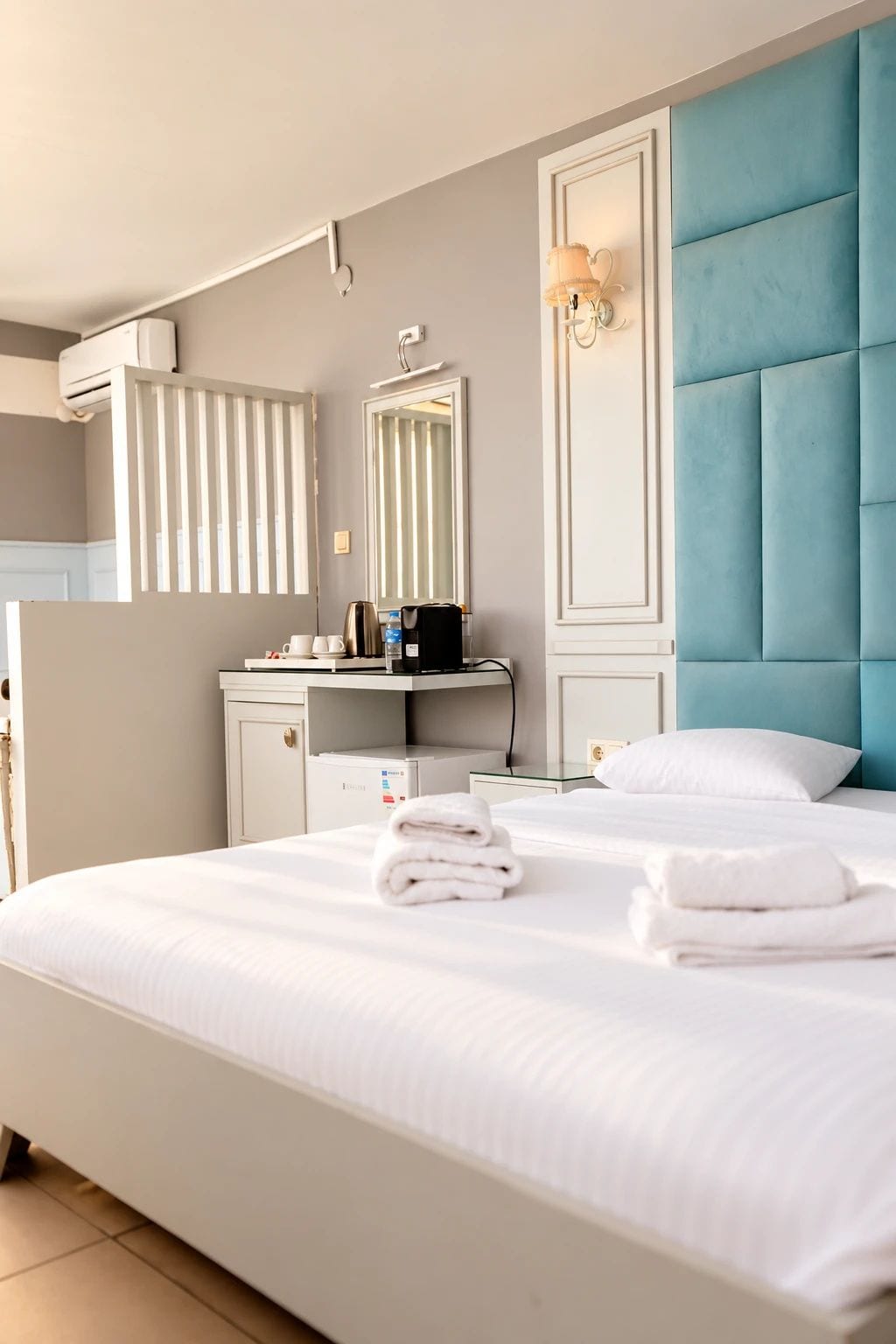 Une chambre d’hôtel comprenant un lit, une coiffeuse avec un miroir, une tête de lit bleue rembourrée et un climatiseur fixé au mur.
