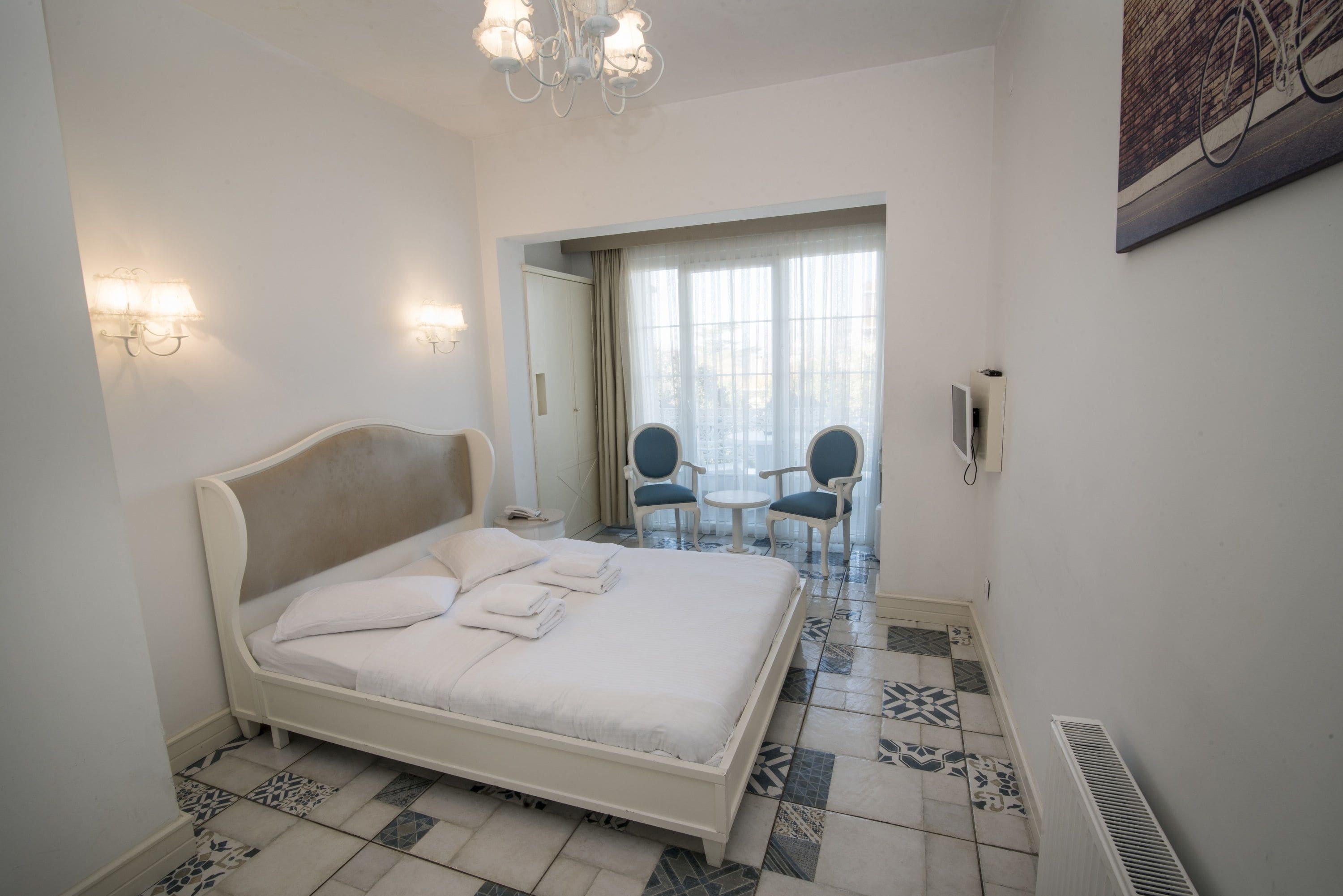 Un dormitorio con una cama grande, dos luces fijadas a la pared, una lámpara de araña, un radiador, un espejo y vistas al exterior a través de una ventana.