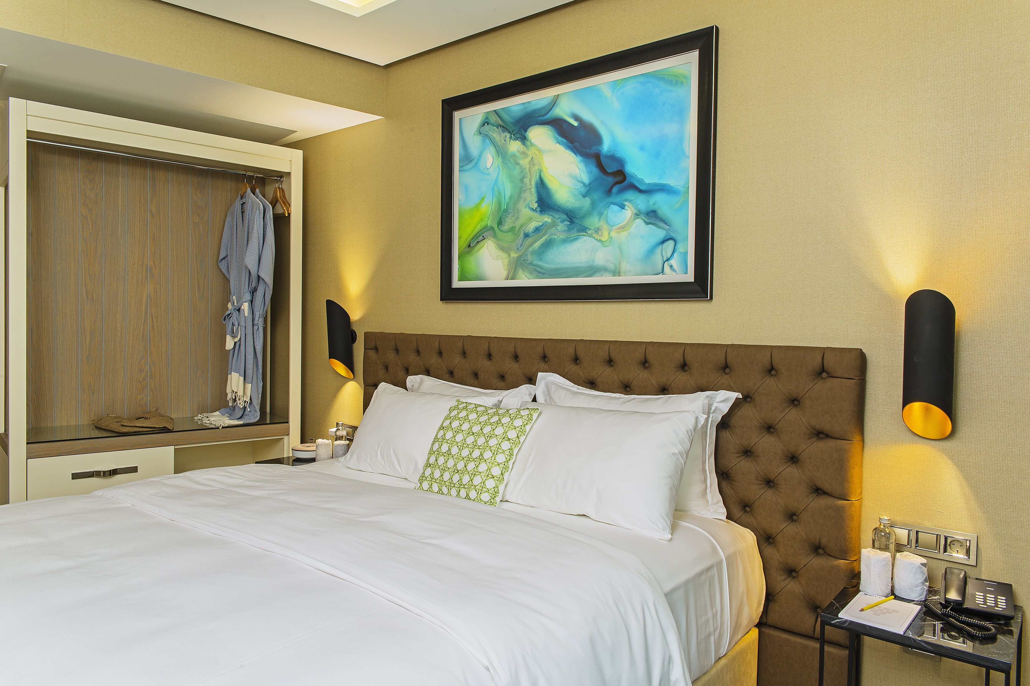 A hotel room with a large bed, a wardrobe, a framed abstract painting, bedside lamps, and a small table with a phone and a bottle.