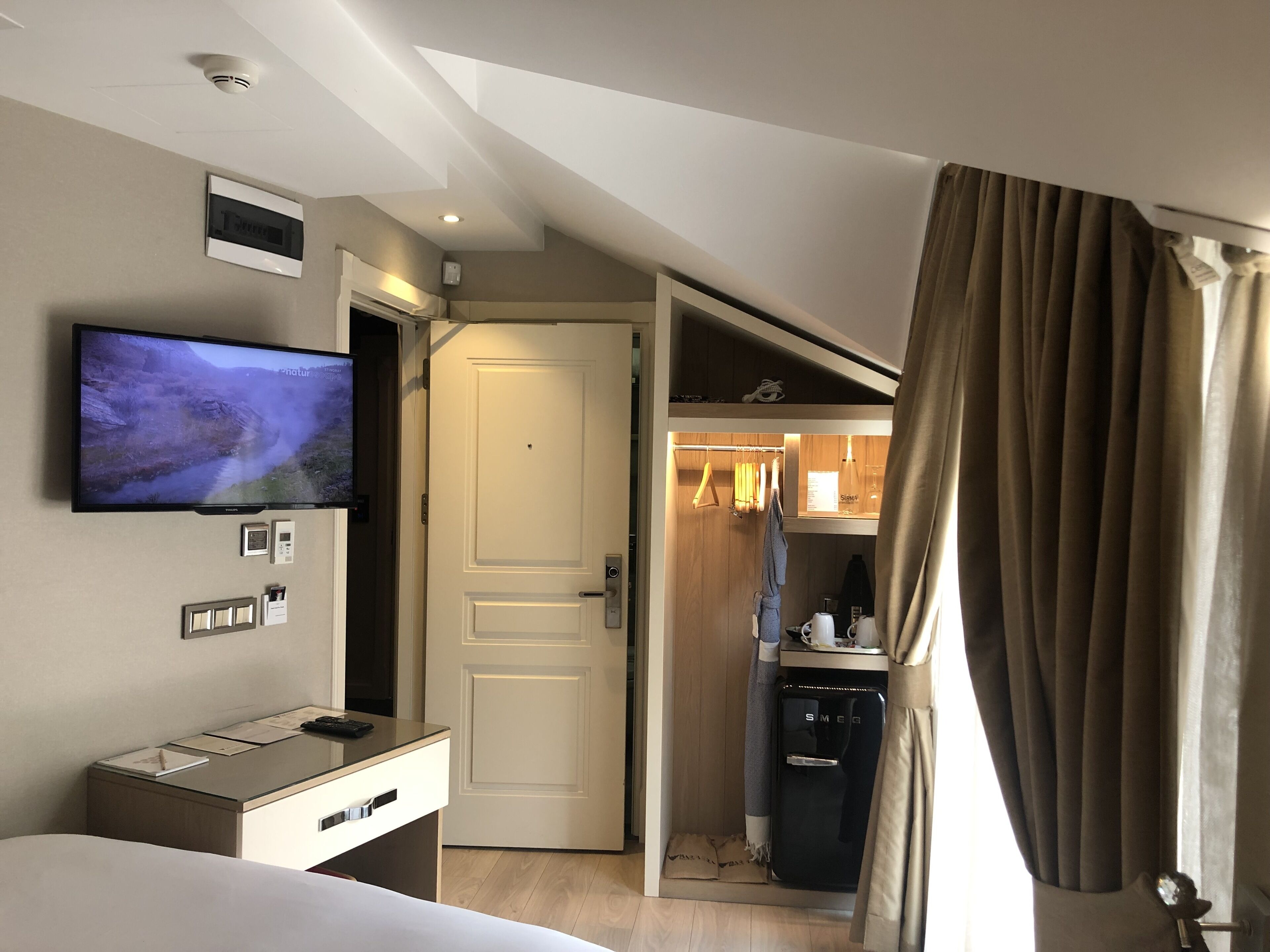 A hotel room with a bed, a TV mounted on the wall, a desk with a computer, a minibar, and a door leading to a bathroom.